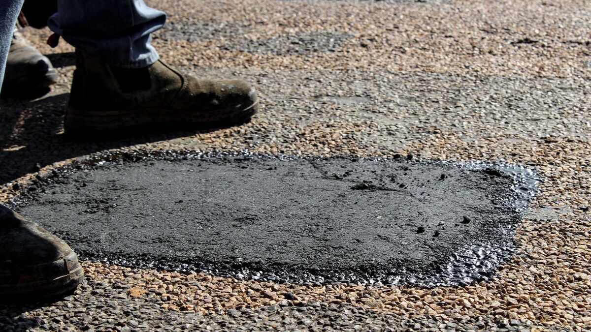 A fresh dark asphalt patch filling a hole in an older, worn pavement surface, with work boots beside it
